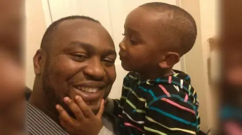 Family Handout Chizi is shown being held by his father in their home. He is wearing a black top with colourful stripes. His father is wearing a striped shirt and is smiling. Chizi is about to kiss his dad on the cheek. 