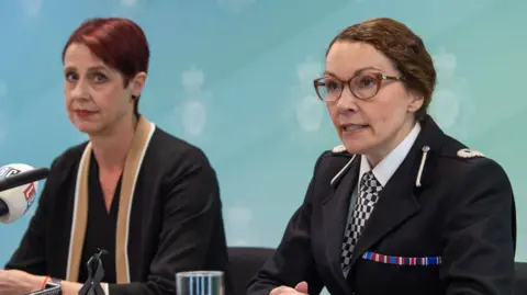 PA Media Two women are seated in front of microphones with a blue background behind them. The woman on the left is wearing black clothing with a beige trim. She had short, dyed red hair and looks at the camera from the side. The woman on the right is dressed in a police unform and has glasses. She has brown hair and looks off camera towards the gathering of reporters