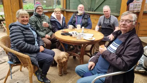 The Drovers Return Cafe Six people, all sitting down, at a table, with mugs of tea and coffees in front of them. A dog is underneath the round table. One man has a hat on, the rest are wearing coats, with one man in short sleeves and an apron. They are in a cafe, they looks like a wooden shed. 