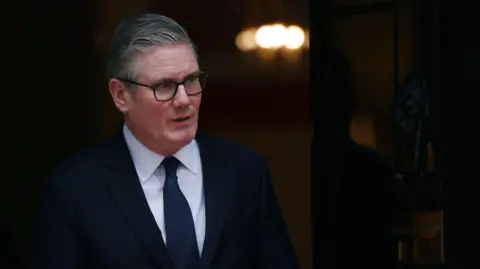 EPA Sir Keir Starmer looking out of a door to the right of the image. He is wearing a dark suit, blue tie and white shirt, as well as glasses. A light can be seen in the background but it is out of focus.