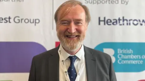 Hampshire Chamber of Commerce Picture shows man in suit with beard smiling at camera with information board behind him