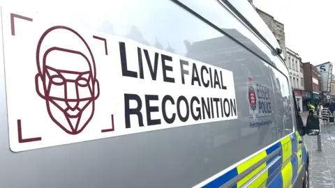 A close up of a grey police van parked up. It has the words live facial recognition written on it as well as the Essex Police logo.