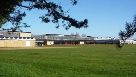 External view of Smithdon High School in Hunstanton. A two-storey building with lots of glass windows. In front of it is a green grass hockey pitch. 