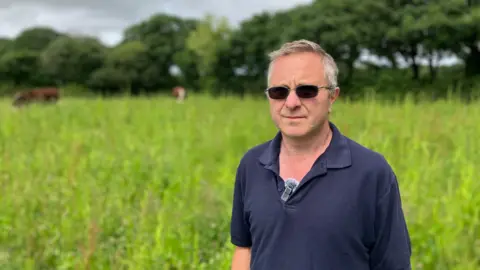 A farmer in a blue short sleeved shirt stands in a large field. The plants in the field are waist high and there are 2 bulls in the background. The sky is overcast.