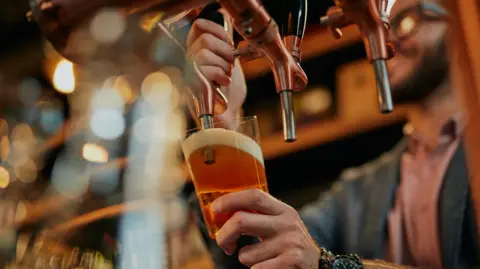 Getty Images A man with a black beard and square glasses pulls a pint of lager. The glass is tilted underneath the pump and the beer has a foamy, white head. There are two other unused beer taps.