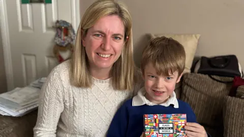 A boy wearing a blue school jumper and a white top sits on his mother's lap on a sofa in a living room. The boy has light brown hair that rests over his forehead. He is holding a card covered in world flags and is smiling. His mum has shoulder-length blonde hair and is wearing a white jumper.