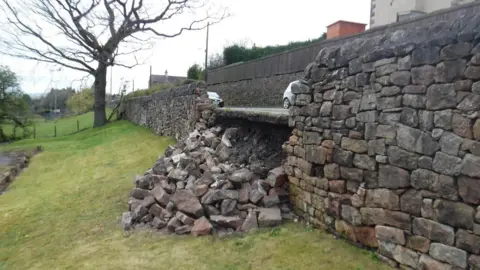 Part of a stone wall running along a road has collapsed on to the grass beneath it. Cars can be seen driving along the road in the background.