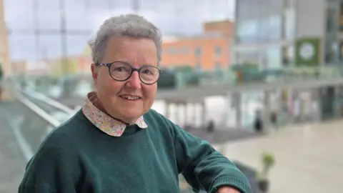 Catherine Rowett is leaning on a balcony on a glass-fronted building. She's wearing a green jumper over a multicoloured patterned shirt.