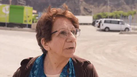 An elderly woman wearing glasses stands talking to camera in a tarmacked area with some vehicles in the background 