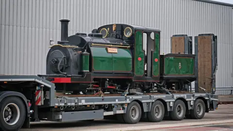 Ian Castledine A vintage green steam locomotive named "Prince" is loaded on a flatbed trailer parked beside a large industrial building 