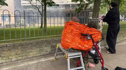 A woman and a child sit on a mental bench and a walking aid covered in a orange blanket. A man can be seen looking over to brown concrete building surrounded by smoke.