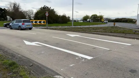 A roundabout at Canvey Island, with two lanes of traffic approaching it and other vehicles already on the roundabout. The left-hand lane shows a left-turn only in white paint, whilst the right-hand lane shows straight on.