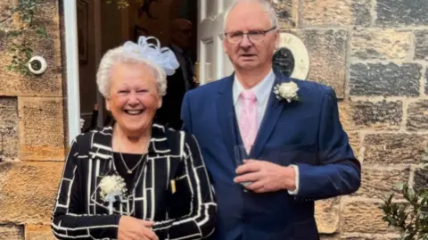 Handout Hubert and Maureen Dodds standing in front of a stone house. They look like they're dressed for a wedding with Mrs Dodds on the left wearing a white fascinator, black and white top and a white rose attached to her right. She is smiling into the camera. Mr Dodds is on the right and in a blue suit with pink tie. He is holding a glass in his hand and has a white rose attached to his lapel. He is looking to the left of the camera.