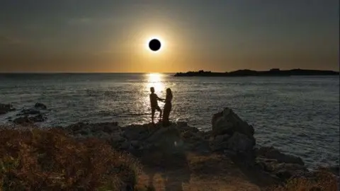 The ark shape of the moon is in front of the sun, whose rays are shining out across the sea. The silhouettes of a couple are seen on rocks on the headland with the sea behind them and the sun's rays reflected in the water.