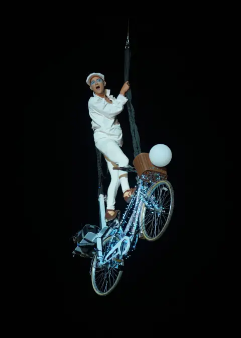 Yui Mok/PA Media A man in white clothing stands on a lit-up bike, which is suspended in the air.