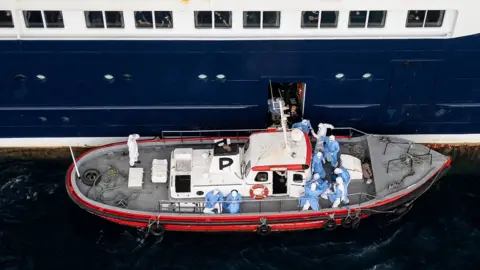 Getty Images Aerial view shows health personnel assisting patients onto a boat from the cruise ship MV Hondius, while stationary off the port of Praia, the capital of Cape Verde.