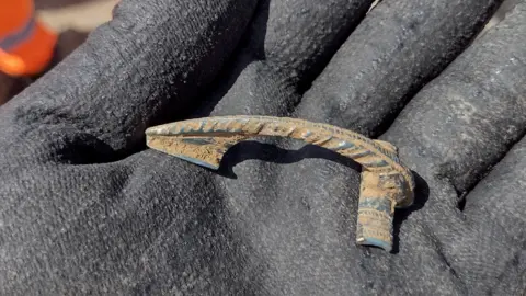 National Highways A gloved hand holding a Roman artefact found in the A417 excavation.