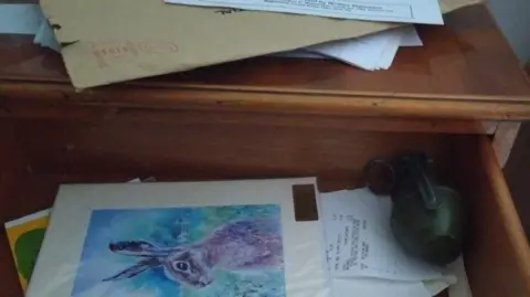 Contributed Hand grenade in the back right-hand corner of an open drawer on top of some pieces of paper and next to a greetings card. Envelopes and letters are on top of the chest of drawers.