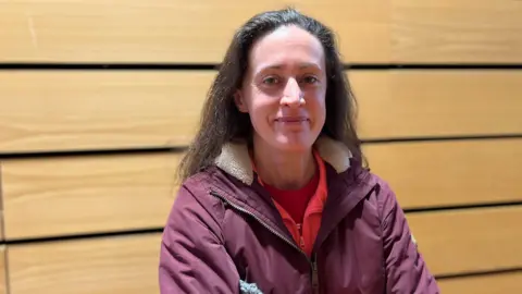 A woman with long dark hair, red top and purple jacket standing in front of a wood-panelled wall