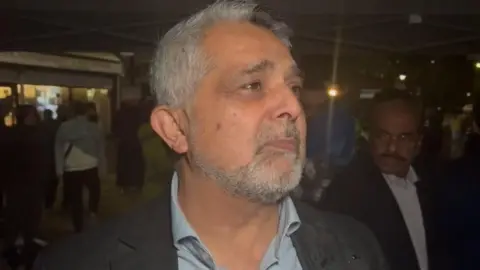 A middle aged man with grey hair and beard speaks to a reporter. It is night time and there are people in the background. 
