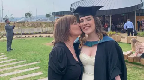 Sophie Morgan (right) pictured with her mother Erica in her cap and gown for her graduation. She smiles at the camera while her mother kisses her cheek. They are both stood outside. 