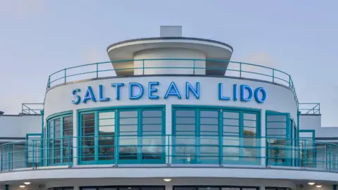 The upper floor of Saltdean Lido seen from the outside with neon letters and an art-deco design. It has a curved window and balcony with frames and railings painted in turquoise.