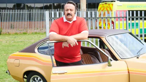 BBC Studios Mike Bubbins stood next to a yellow car. He is leaning on the window frame wearing red polo shirt