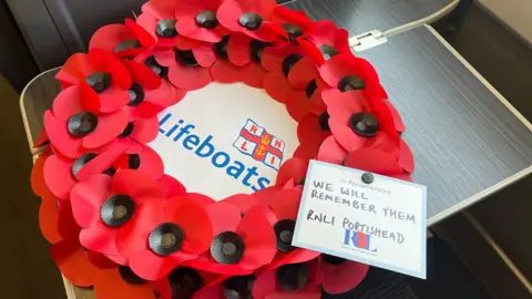 A poppy wreath laid out on a table on a Great Western Railway train. In the middle of the wreath is a white piece of paper with the RNLI logo and the word 'lifeboats' written in blue font. There is a white piece of paper attached to the wreath which says "In remembrance. We will remember them. RNLI Portishead.