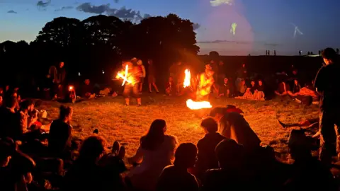 A circle of people at night all watching two people in the middle perform tricks with fire to mark the summer solstice in Wiltshire