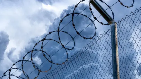 Getty Images Close-up of metal fence with barbed wire over a stormy blue sky