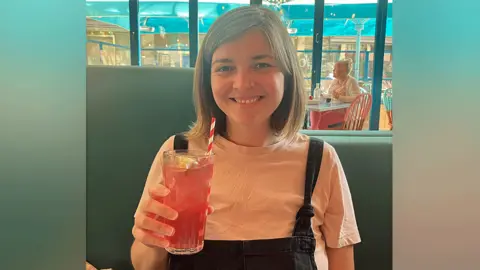 Lizzy Willmington Lizzy Willmington raise a cocktail glass full of a red drink as she smiles for the camera. She is wearing a white t-shirt and black dungarees with her light hair reaching her shoulders. She is sitting on a seat with a blue back, with other diners in the background. 