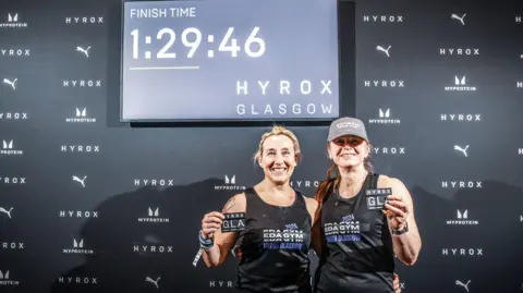 Hyrox and Sportograf Leigh and Wiki hold up their competition badges under a digital display of their finish time. They both are smiling for the camera.