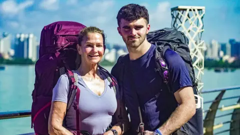 BBC/Studio Lambert Caroline and Tom are stood with a large body of water behind them. Both are wearing t-shirts and wearing huge backpacks.
