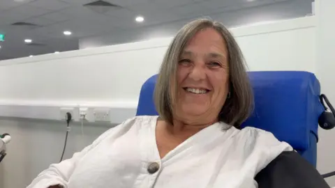 Image shows Sue smiling at the camera. She had short hair. She is wearing a white top with brown buttons on it. She is sat in a dark blue chair. 