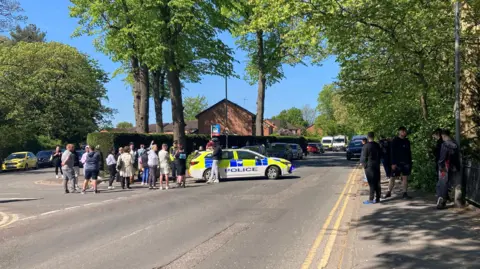 A large number of people are gathered around a police car which is positioned in the middle of the road. There are further police vans in the distance. There are trees either side of the road.