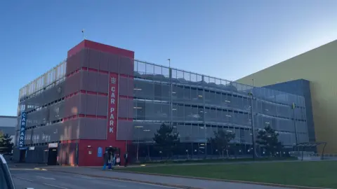 A large multi-storey car park set against a blue sky. The structure is covered in silver lattice-shaped screens. The left part of the building is a large red tower with the word "car park" written on the site. Three green trees and a grassed area are visible in the foreground of the image.