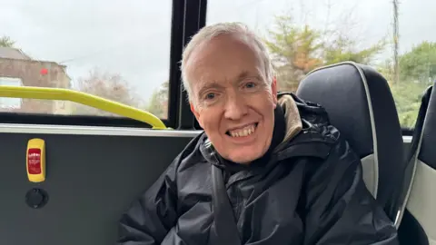 A man with grey hair smiling at the camera. He is sitting on a bus with a seatbelt on. He has a black coat on and the background outside the bus window is grey and gloomy.