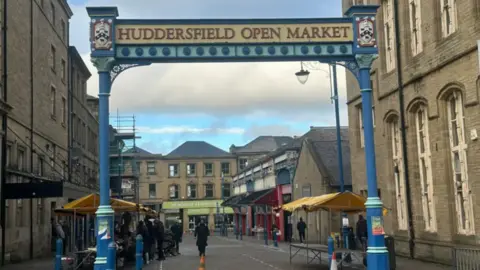 OLIVIA COURTNEY-ASHTON/BBC Shot of the welcome posts to Huddersfield Open Market. They are painted blue and line either side of the street. A banner at the top reads 'Huddersfield Open Market' in red lettering, against a yellow backdrop.