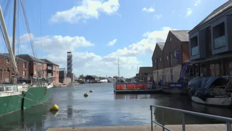 The photo shows Exeter Canal. It is a waterway with industrial-looking buildings on either side. Some boats of different sizes can be seen moored on the water. 