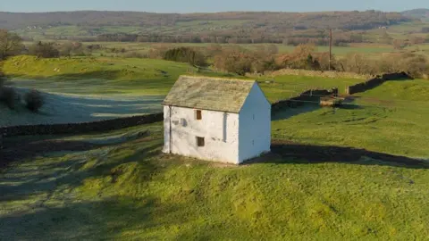 An aerial shot of Henry's Castle. It is a small white building with a pitched roof set in the middle of green countryside. It has two small widows on one side.