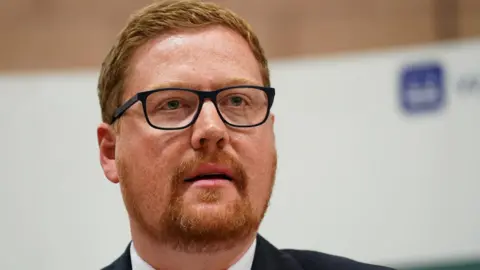 Jonathan Brash MP. He has ginger hair which is closely cut and neat as well as a ginger beard and goatee. He is wearing a pair of black glasses.