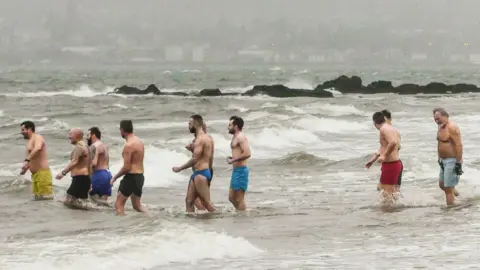 Tommy Bamford Ten swimmers in just shorts up to their knees walking into very choppy water.