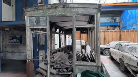 Looking through the wooden shell of the dilapidated tram carriage. There are gaps where the window and doors would have been and a large pile of wooden slats in the interior.