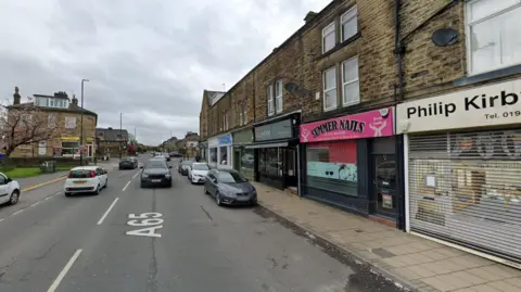 Parade of shops on Otley Road in Guiseley, Leeds. Cherry blossom tree and stone house to the left. Terrace of stone built shops to the right, including Summer Nails (pink sign) and Philip Kirby jewellers (white sign). Cars and A65 Google writing in the road, parked cars on the right.