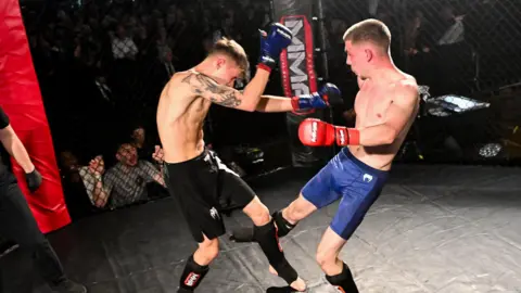 Ultra Events Two young men in a Mixed Martial Arts cage engage in a fight. They both wear shorts, boxing gloves and protective boots.