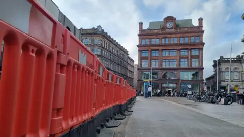 An orange barrier lines a street. A large building with a clock on top is in the background, in front of this is people walking, street signs and a some bikes which are locked up .