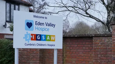 Eden Valley Hospice Close-up image of a sign reading "Welcome to Eden Valley Hospice". Below that is the logo for Jigsaw, Cumbria's Children's Hospice, which is also based at the site. A brick wall can be seen behind the sign along with a section of a white building.