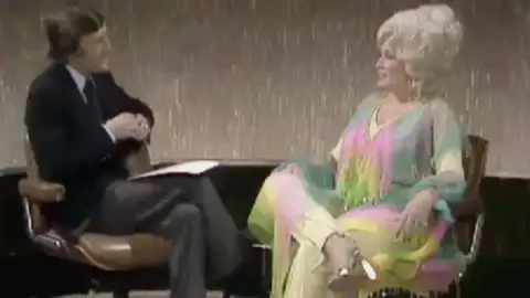 A man in a dark suit interviewing a woman in a colourful outfit.