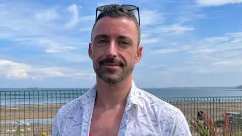 A man with short brown hair, wearing a red top and a white short sleeved shirt over it. He is standing in front of a beach.