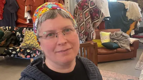 Mousumi Bakshi/BBC Julie Smith is to the left of the image. She has short grey hair and is wearing a patterned head band. She is wearing a black T-shirt and a grey jacket, and is standing in front of two sofas which have fabrics resting on them.